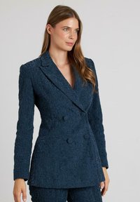 Blazer de doble botonadura en tejido texturizado en verde azulado con un patrón floral, solapas de muesca y un ajuste a medida.