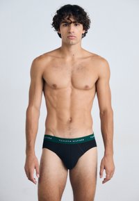 Schwarze Baumwollslips mit einem gebrandeten Bund in Dunkelgrün, mit klassischem Schnitt und glatter Textur. Das gezeigte Modell steht aufrecht.