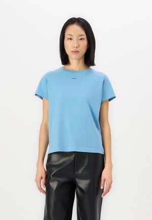 Helles blaues Baumwoll-T-Shirt mit kurzen Ärmeln und rundem Halsausschnitt. Mit einem kleinen Aufdrucklabel auf der Vorderseite. Kombiniert mit schwarzen Lederhosen.