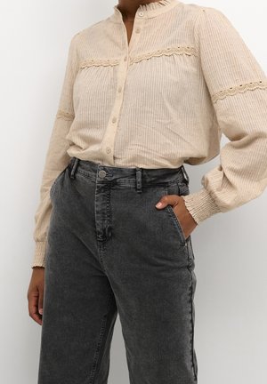 Beige bluse med lange ærmer og blondedetaljer, båret med højtaljede falmede sorte jeans, modellens højre hånd i lommen mod hvid baggrund.