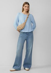Hellblaues Langarmoberteil mit gerafftem Ausschnitt, kombiniert mit weiten blauen Jeans. Die Accessoires umfassen eine strukturierte beige Umhängetasche.