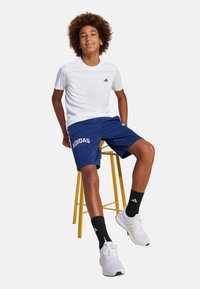 Camiseta deportiva blanca y pantalones cortos navy con el texto "ADIDAS", combinados con calcetines negros y zapatillas blancas. Sentado en un taburete amarillo.
