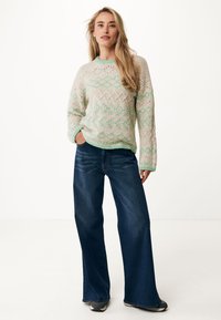 Stickad krämfärgad tröja med mintgrön geometrisk mönstring, matchad med mörkblå vida jeans. Bakgrunden är enkel.