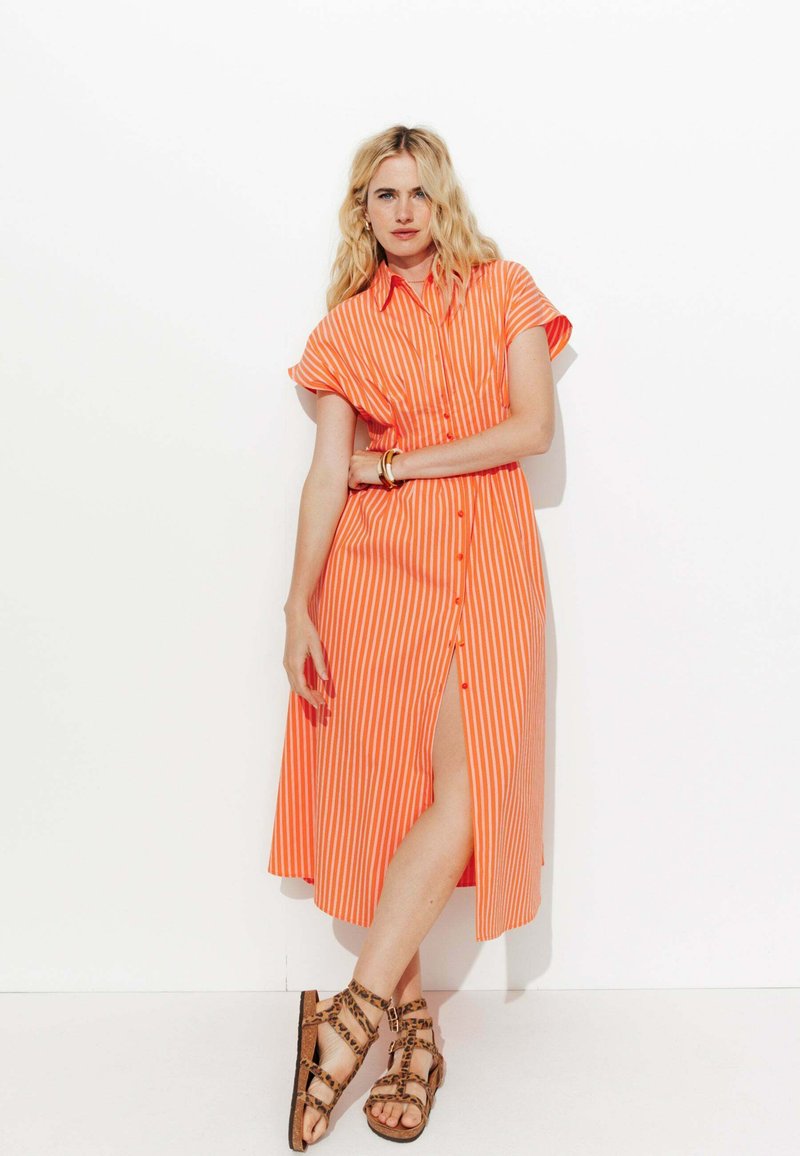 Robe chemise à rayures orange et blanches avec des manches courtes, fermeture à boutons sur le devant et fente sur le côté. Comprend des sandales marron à lanières.