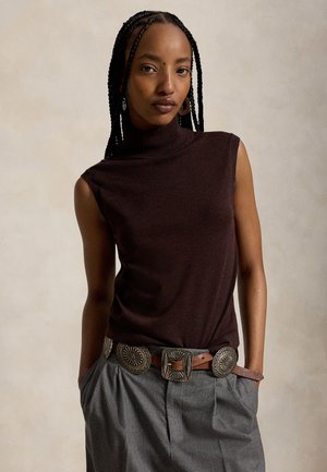 Femme aux cheveux tressés portant un haut marron sans manches, pantalon gris taille haute avec une ceinture marron décorative, mains dans les poches, fond neutre.