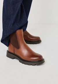 Brown leather Chelsea boots with elastic side panels, round toe, raised ridged sole, and contrasting black trim, paired with dark denim trousers.
