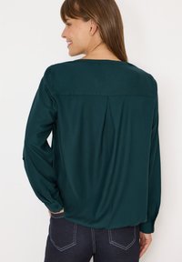 Blouse à manches longues en teal avec un pli dans le dos, des poignets boutonnés et une texture lisse. Portée avec un jean foncé présentant des coutures contrastantes.