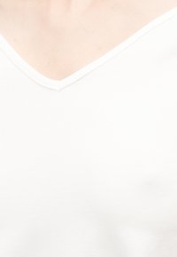 Gros plan sur une personne portant un t-shirt blanc uni à col en V, mettant l'accent sur l'encolure et la partie supérieure de la poitrine.