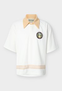 Λευκό πόλο μπλουζάκι με κοντά μανίκια, μπεζ μυτερό γιακά, μπεζ ριγέ διακοσμητική ταινία στο τελείωμα και κυκλικό έμβλημα "HEATHE Football Federation 1978" στο στήθος.