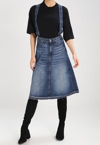 Kvinna iförd svart topp, knä-lång blå denimkjol med hängslen och svarta knähöga stövlar mot en enkel ljus bakgrund.