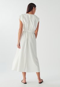 Bílá, bezrukávová šaty se staženým pasem, splývavou sukní a jemným zapínáním na knoflíky na zadní straně, vyrobené z lehkého materiálu.