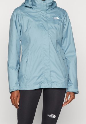 Chaqueta impermeable azul claro con capucha y bolsillos con cremallera, usada sobre leggings negros, ambos con el logo de The North Face.