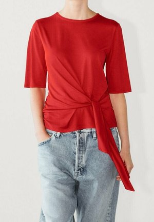Haut rouge à manches courtes avec un détail torsadé à la taille, fabriqué en tissu lisse. Associé à un jean taille haute bleu clair.