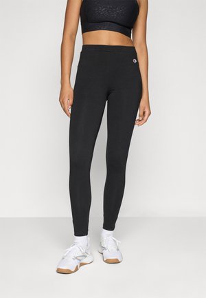 Woman wearing black high-waist leggings, black crop top, white socks, and white sneakers standing against a plain light background.
