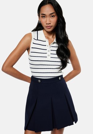 Woman with long black hair wearing a sleeveless white polo top with black stripes and a navy pleated skirt with button details.