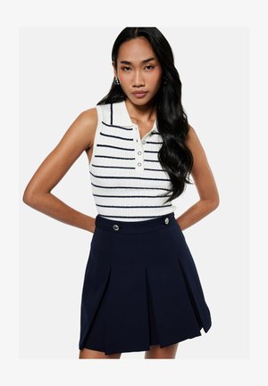 Frau mit langen schwarzen Haaren, die ein ärmelloses weißes Poloshirt mit schwarzen Streifen und einen marineblauen Faltenrock mit Knopfdetails trägt.