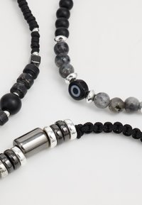 Collane di perline con perle nere opache, pietre grigie macchiate, accenti argentati e corda nera intrecciata. Texture e finiture diverse.