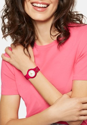 Montre en silicone fuchsia avec un cadran rond blanc, des accents dorés et un charme en forme de cœur, associée à un t-shirt rose.