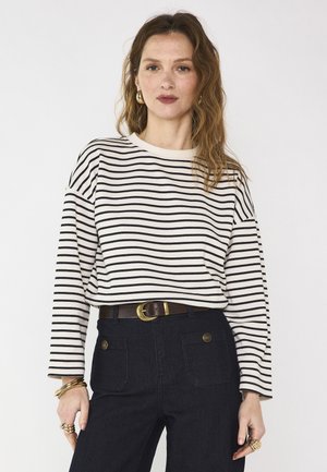 Frau mit welligem Haar, die ein weiß-schwarz gestreiftes Langarmshirt, dunkle, hoch taillierte Hosen mit Fronttaschen, goldene Ohrringe und Ringe trägt.