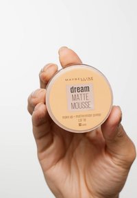 Maybelline Dream Matte Mousse in un contenitore rotondo e trasparente con un'etichetta color pesca. La consistenza sembra liscia, etichettata "30 sabbia."
