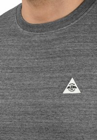 T-shirt grigia con texture, girocollo, con un piccolo etichetta triangolare bianca con lettering nero sul lato sinistro vicino al colletto.