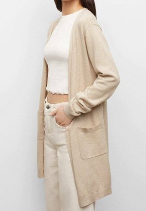 Frau trägt einen beigen langen Cardigan über einem weißen, schmal geschnittenen, gerippten Crop-Top und hellen, hochtaillierten Hosen, Hand in der Tasche, vor einfachem Hintergrund.