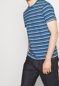 Randig blå t-shirt med horisontella vit- och mörkblå ränder, med en liten röd logotyp på bröstet. Materialet verkar mjukt och andningsbart.