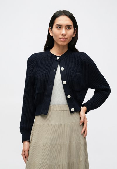 Μπλε κασμιρένια ζακέτα με ριμπ υφή, στρογγυλή λαιμόκοψη και μεγάλα εκρού κουμπιά. Διαθέτει δύο μπροστινές τσέπες και κοντό μήκος.