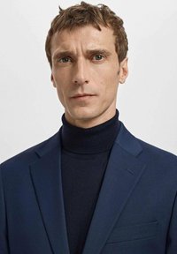 Marineblauer Blazer aus strukturiertem Stoff, getragen über einem dunklen Rollkragenpullover. Der Blazer verfügt über scharfe Revers und eine einzelne Vorderstasche.