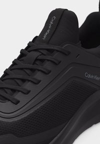 Κοντινό πλάνο μαύρου αθλητικού παπουτσιού Calvin Klein με υφή από δίχτυ, σχέδιο με κορδόνια και λογότυπο στη γλώσσα και στην πλευρά.