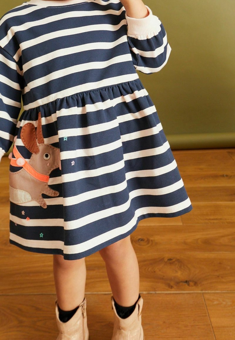 Enfant portant une robe rayée bleu marine et blanche avec un patch lapin marron, debout sur un sol en bois, bottes beiges visibles.