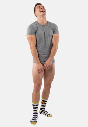 Giovane uomo con maglietta grigia e calzini a righe a tema spaziale che ride, coprendo le gambe nude con le mani su uno sfondo grigio chiaro.