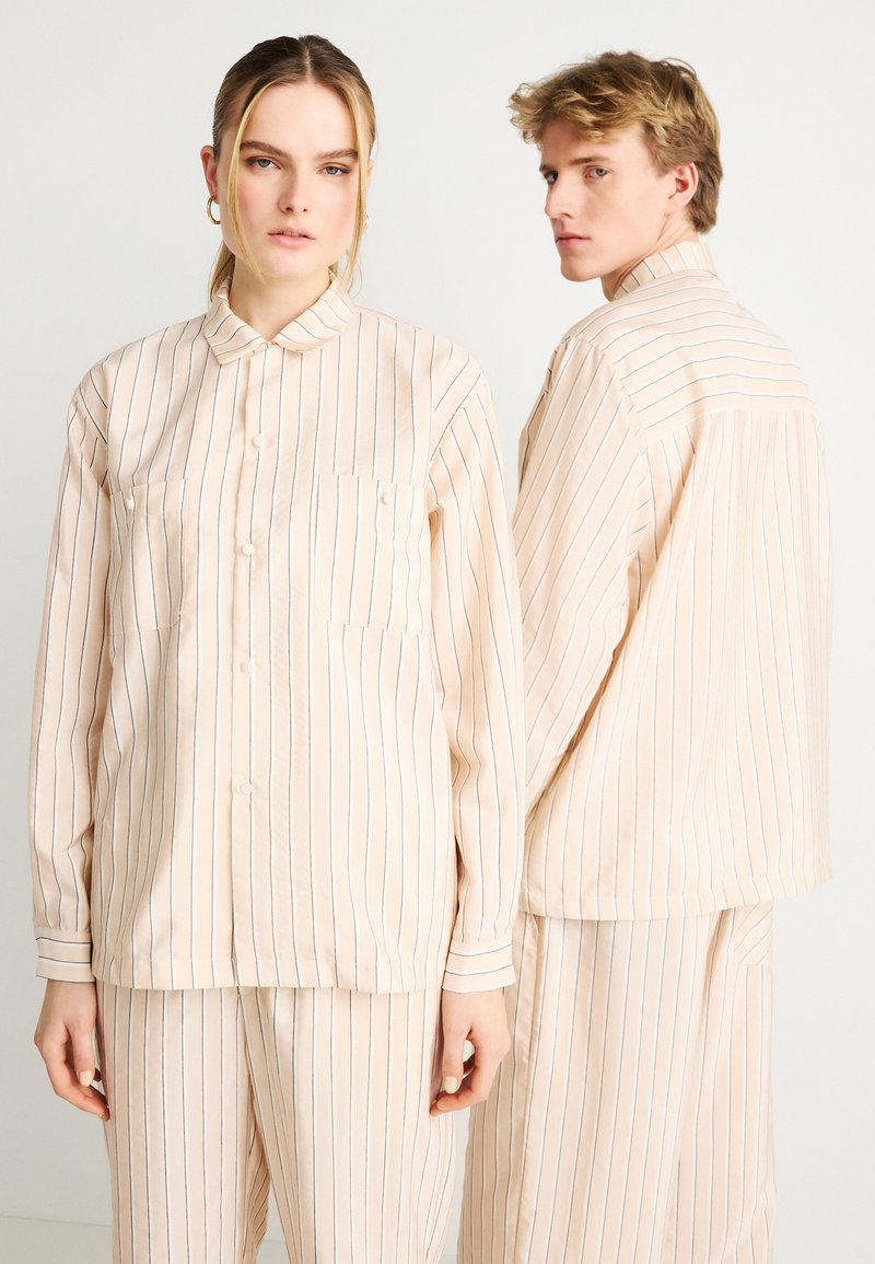 Σετ πιτζάμες σε ανοιχτό ροδακινί χρώμα, με κάθετες μπλε ρίγες. Το πουκάμισο έχει γιακά, κουμπιά και μπροστινή τσέπη· το παντελόνι διαθέτει ελαστική μέση.