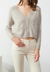 Cárdigan corto de un beige claro con escote en V, bordes acanalados y tres botones decorativos; textura suave y dos bolsillos frontales. Combinado con pantalones de color claro.