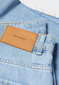 Vaaleansiniset farkut, joissa on tan-värinen nahkapatja, johon on ommeltu "MNG DENIM" -merkintä taka-taskun yläpuolelle ja näkyvä vyölenkki.