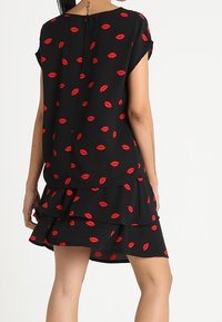 Mujer vestida con un vestido negro de manga corta con un patrón de labios rojos, falda de volantes en capas y cierre en la parte posterior, de pie frente a un fondo blanco.