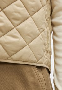 Colete bege acolchoado com padrão de diamante, apresentando uma superfície texturizada e uma etiqueta com a inscrição "Marc O'Polo." Usado por cima de uma blusa creme clara.