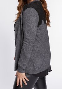 Šedý kardigan s krajkovým detailem na ramenech, dlouhými rukávy a volnějším střihem. Texturovaná látka zvýrazňuje kontrast s hladkou krajkou.