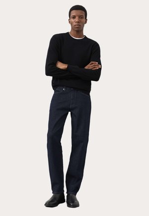 Mand stående med armene over kors iført en sort sweater, mørkeblå jeans og sorte sko mod en ensfarvet lys baggrund.