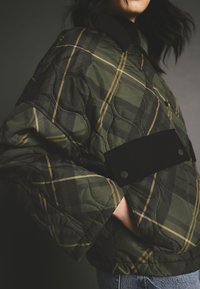 Jachetă cu model pătrățos verde, matlasată, cu accente negre din catifea și buzunare mari cu patch-uri. Are guler și o croială lejeră.
