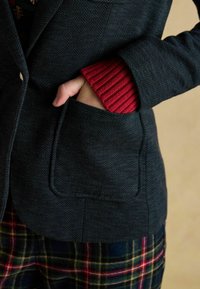 Tmavý sako s rybia kostra vzorom a jednou vreckom; červený pletený manžetový náplet vyčnieva spod rukáva, skombinované s multikolórovými kockovanými nohavicami.