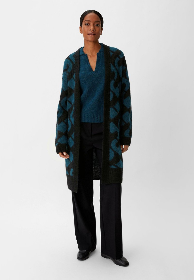 Langer schwarzer Cardigan mit türkisem geometrischem Muster, getragen über einem passenden türkisen Pullover und schwarzen Hosen. Glatte Textur und entspannter Schnitt.