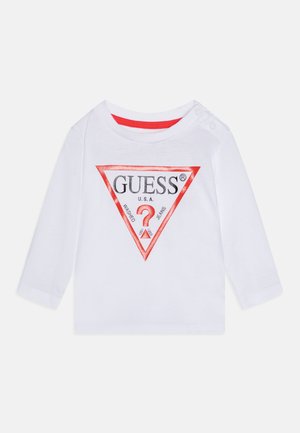 La camisa blanca de mangas largas presenta un logotipo triangular de "GUESS" en rojo y negro en la parte frontal, con botones de presión en el hombro izquierdo.