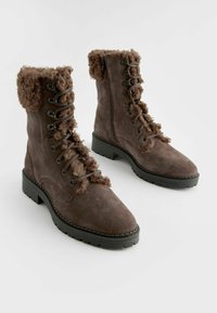 Botas de tobillo de ante marrón con un puño de borrego marrón y esponjoso. Tienen un cierre con cordones en la parte delantera y una suela gruesa y texturizada en color negro.