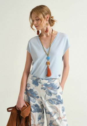 Donna con top azzurro a scollo a V e pantaloni bianchi con stampa floreale blu, che indossa orecchini lunghi con nappine e collana, mentre tiene una borsa marrone con frange.