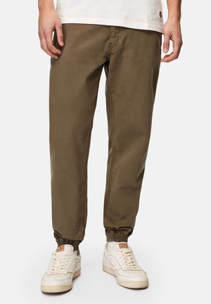 Mężczyzna ubrany w oliwkowe spodnie dresowe z elastycznymi ściągaczami i białe trampki, stojący na jednolitym tle.