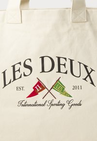 Beige canvas tote bag featuring black text, "LES DEUX," and red and green crossed flags with "H" and "LES DEUX" embroidered.