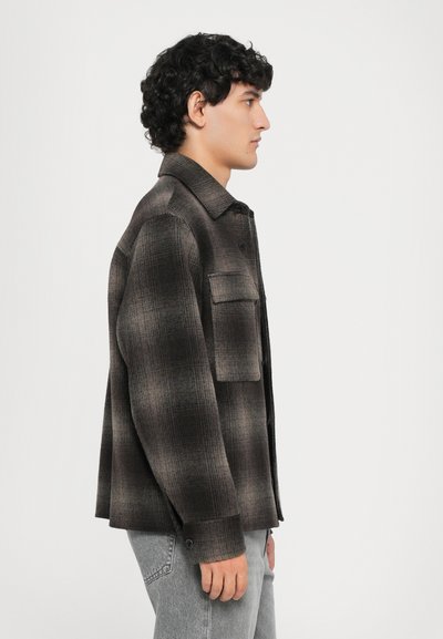 Young man with curly hair wearing a dark plaid button-up jacket and light grey jeans, standing sideways against a plain background.