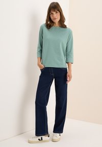 Femme debout, la main dans la poche, portant un haut à rayures verticales vert clair à manches longues, un jean large bleu foncé et des baskets blanches.