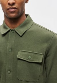 Camicia verde oliva a maniche lunghe in tessuto strutturato, con colletto, chiusura frontale con bottoni e una singola tasca sul petto.